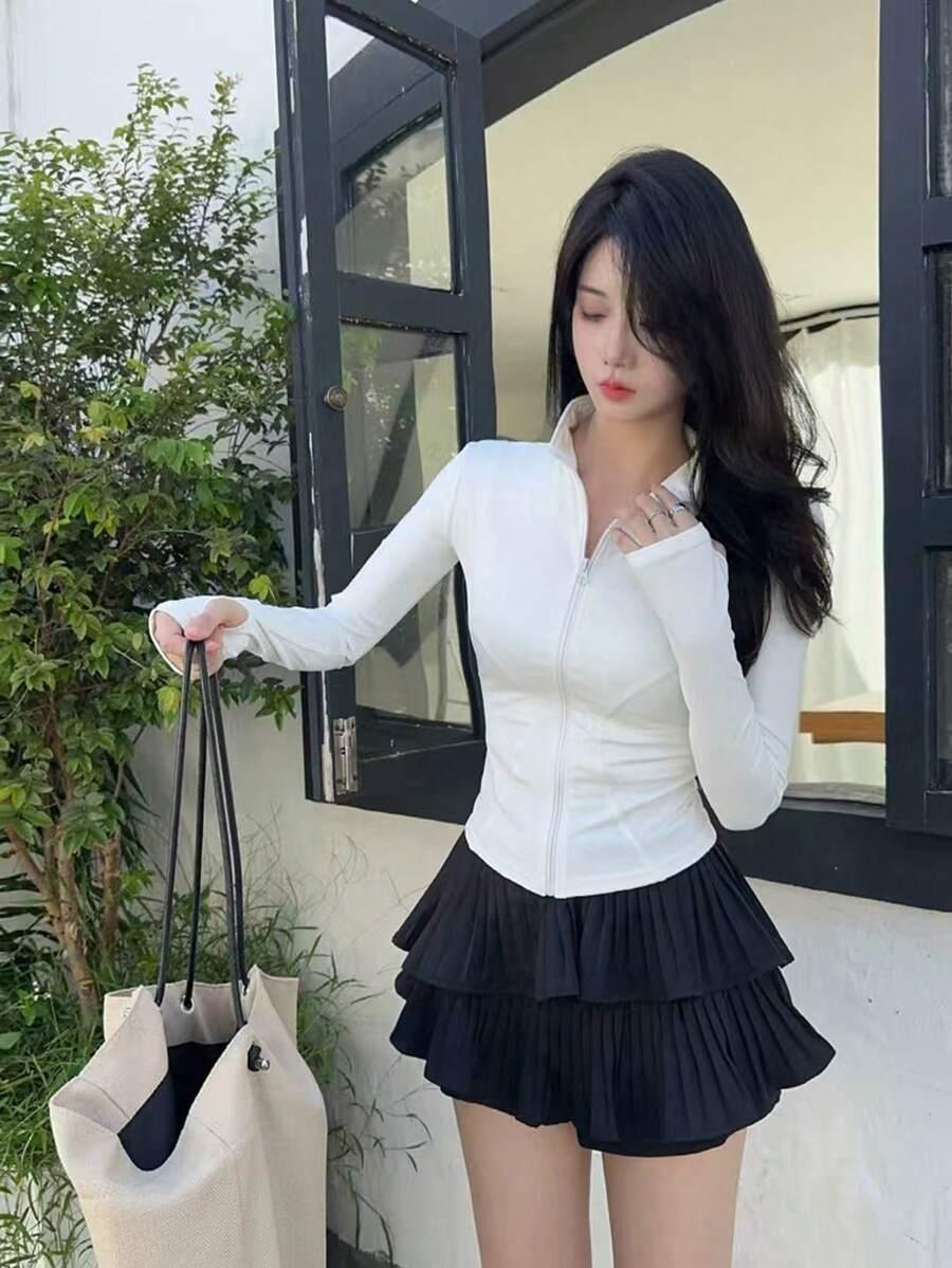 Mulher com jaqueta branca de manga longa e saia preta plissada em fundo branco fosco