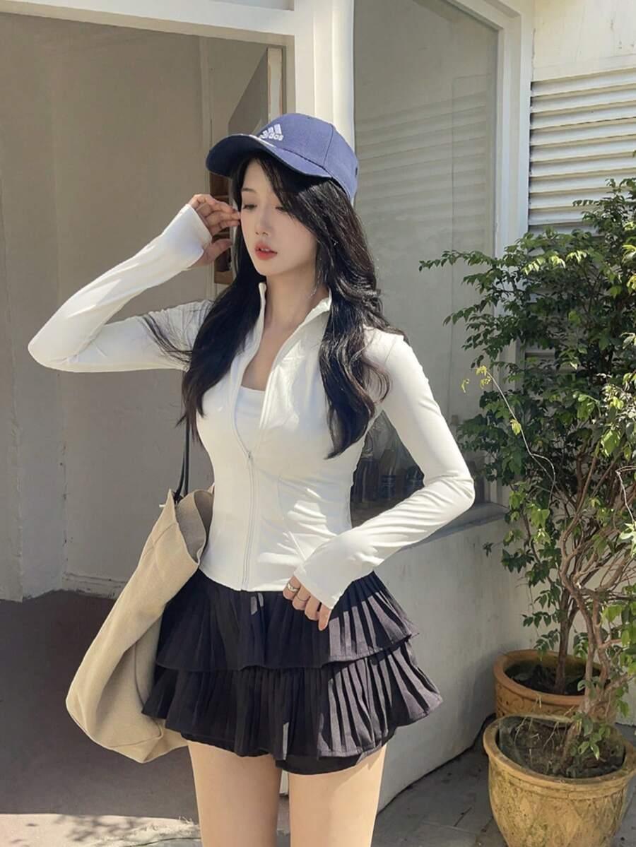 Mulher vestindo jaqueta branca ajustada com zíper, saia preta plissada de camadas, boné azul, fundo branco fosco