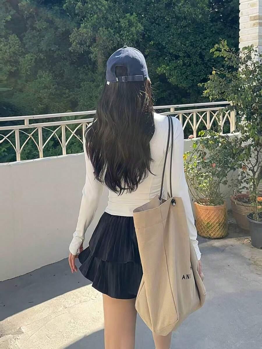 Mulher de costas com blusa branca, saia preta plissada, boné azul e bolsa bege em fundo branco fosco