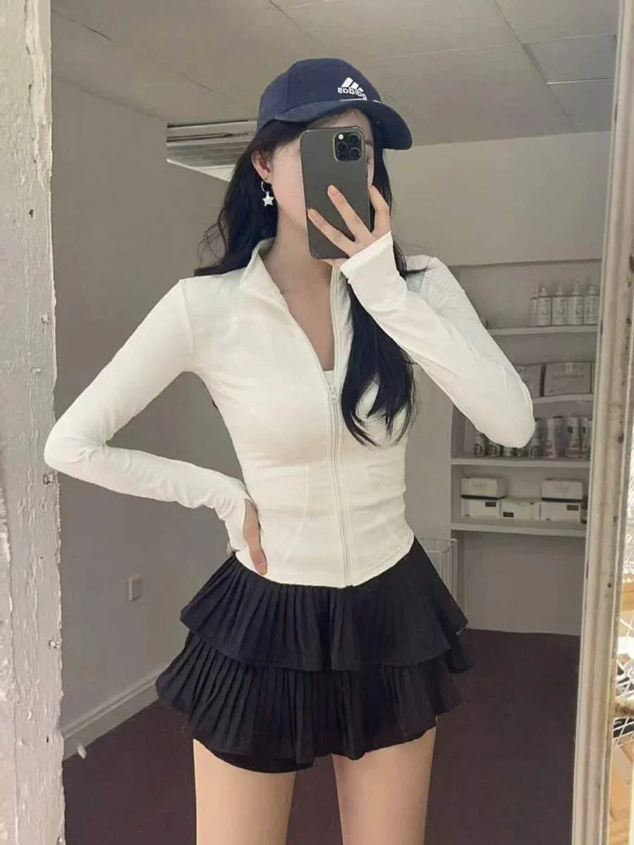 Mulher vestindo jaqueta branca com zíper, saia preta plissada, boné azul, em fundo branco fosco